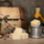 A treasure-themed setup showcasing four artisanal marshmallows with a golden topping and sprinkled oats. In the background, a vintage-style map-covered treasure chest overflows with pearl necklaces, rings, and a jeweled cross. A pewter cup holds one marshmallow, and a bottle wrapped with orange twine adds a rustic pirate vibe.