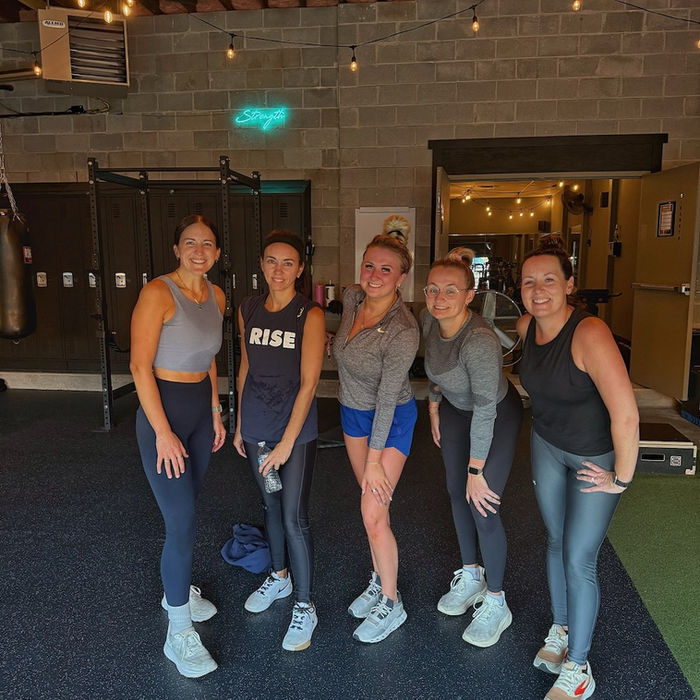 Five women in athletic wear smiling in a gym
