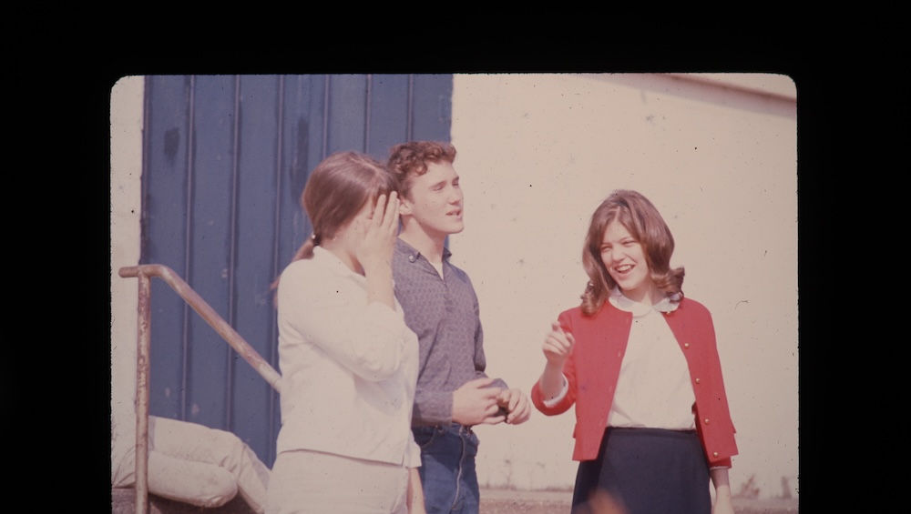 Three young adults smiling, talking casually outdoors, 1960s era.