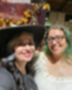 Two smiling women pose together in a brightly lit setting. The woman on the left has long, dark hair with gray streaks, wears a wide-brimmed black hat and black outfit, giving off a witchy vibe. The woman on the right has green-tinted hair, glasses, and a ruffled off-shoulder white top. Autumn leaves and Halloween decorations can be seen in the background.