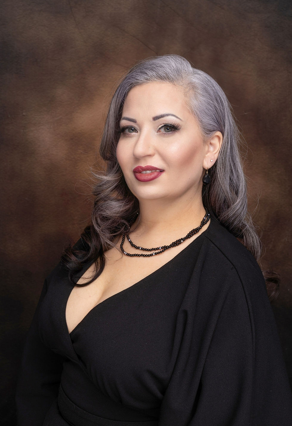 A woman with long, silver-streaked hair styled in soft waves poses confidently against a warm brown backdrop. She wears a black V-neck top, layered black beaded necklaces, and matching earrings. Her makeup is elegant, with bold eyeliner and deep red lipstick, creating a glamorous and sophisticated look.