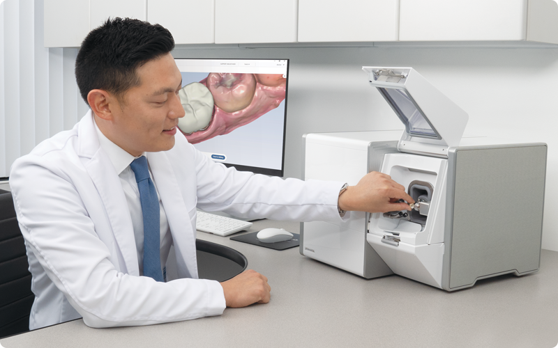 A dentist operates a dental machine, wearing a white coat and blue tie. A dental model is visible on a screen in a clinical setting.