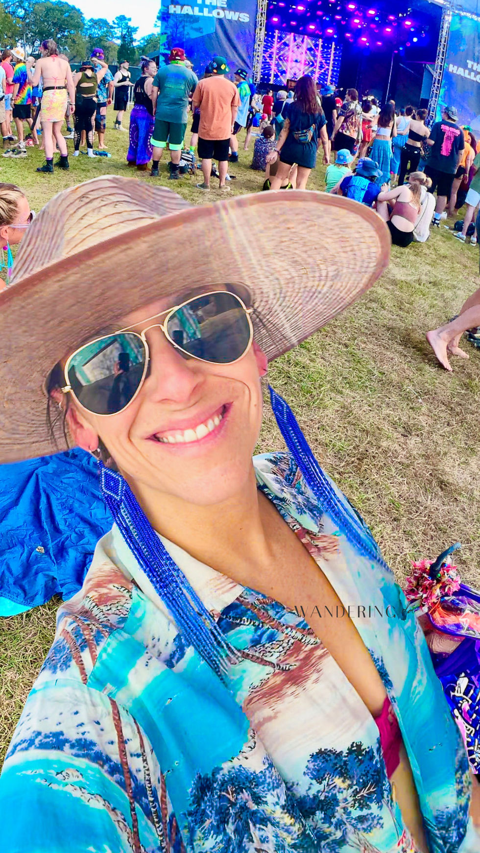 A women wearing handmade beaded fringe earrings at a music festival. She is having a great time, smiling. She is wearing aviator sunglasses, wide brim hat, and a hawaiin shirt.