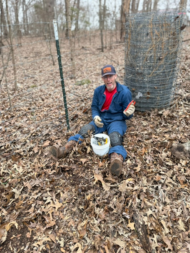 A male with a hat sitting on a bunch of leaves with a fencing tool in his hand and a roll of fencing behind him. He is working on repairing a fence
