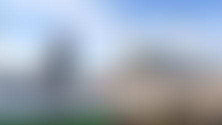 Two people walking in Taksim Square, Istanbul, near a monument and large mosque with minarets under a bright blue sky. Green grass and red plants in the foreground.