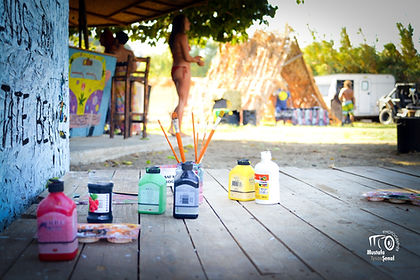 Paints and brushes on wood deck, people in background, camping heavensurfhouse.