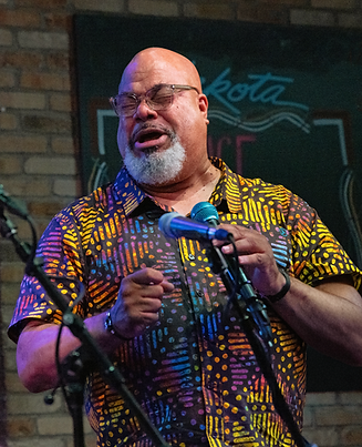 Bald man with grey beard, glasses, and brightly patterned shirt singing at microphone.