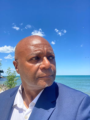 Bald Black man in blue jacket by lake under blue sky