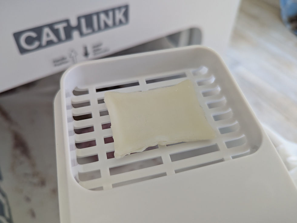 White slotted plastic tray with a deodorizing block in front of a blurred CAT-LINK box, in a clean indoor setting.