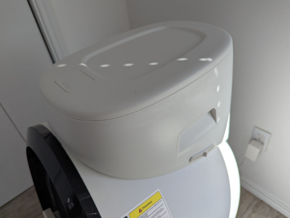 White automatic litter box in a corner by a wall outlet, with sunlight dots and a yellow caution label