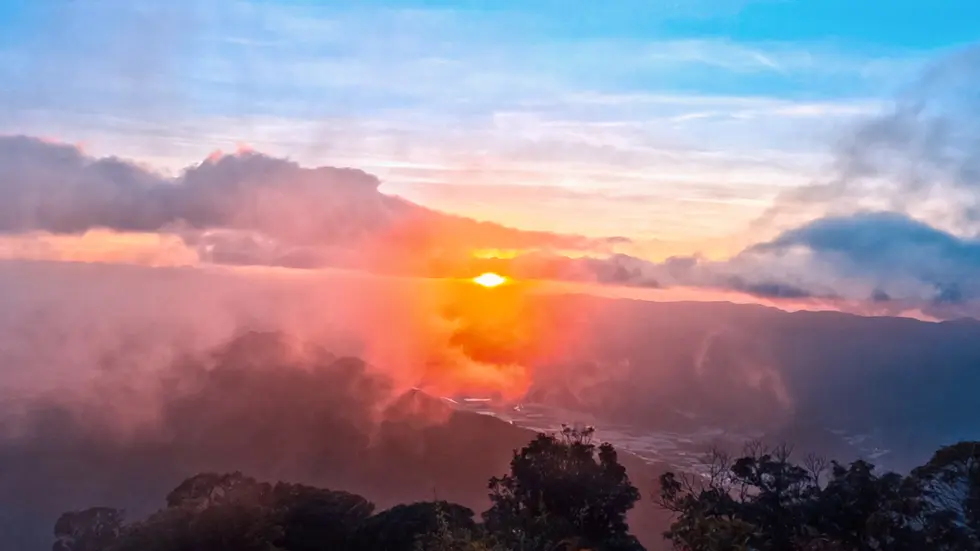 Mist and clouds cover the peaks of a mountain with the orange glow of a sunset behind them surrounded by blue sky in South Vietnam