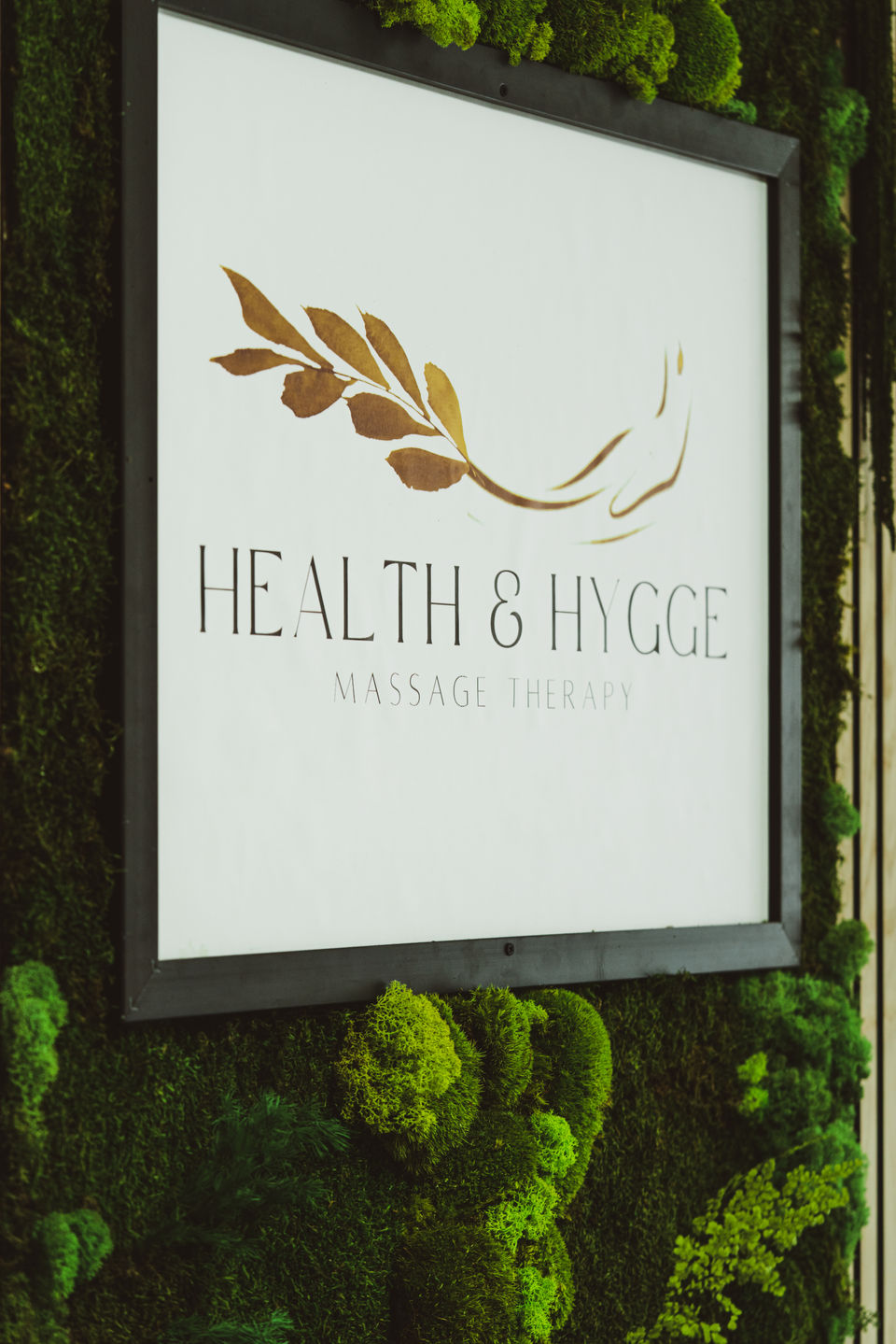 På dette billede kan du se hovedvæggen i Health & Hygge massagestudie. Firmaets logo er trykt på et stort lærred og er omgivet af en dekorativ grøn væg bestående af frodige grønne konserverede mosser, bregner, fyrreblade og andet løv, hvilket giver en fantastisk 3D-effekt til dets naturlige skønhed.