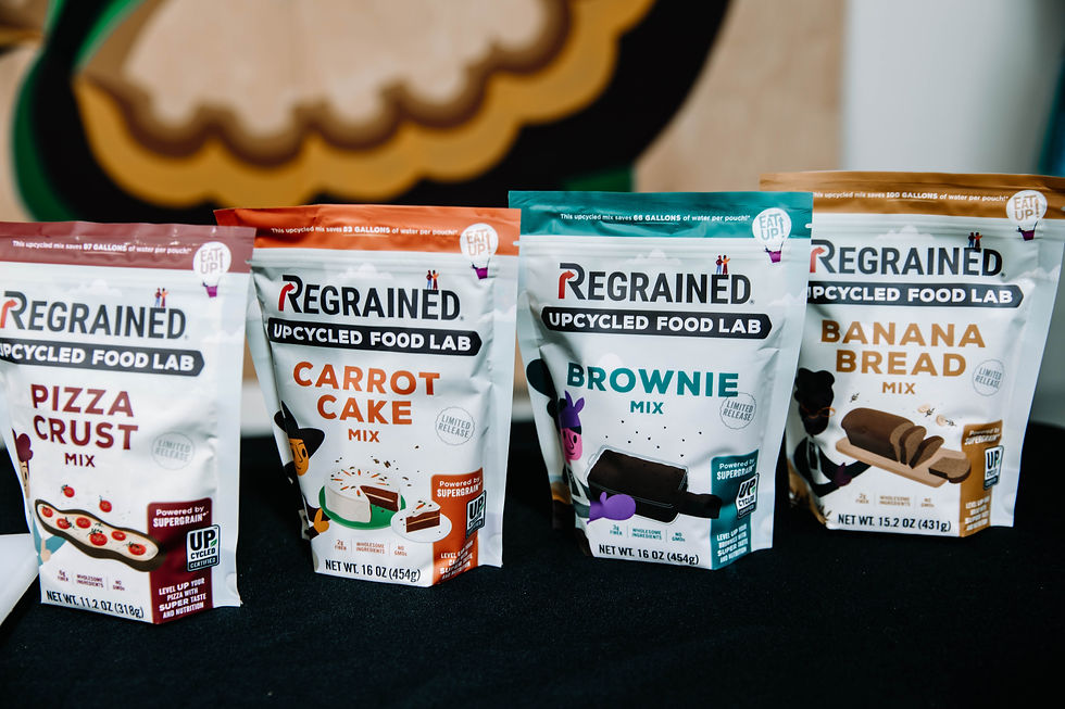 ReGrained Upcycled Food Lab mixes on display: Pizza Crust, Carrot Cake, Brownie, and Banana Bread. Colorful packaging on a black table.