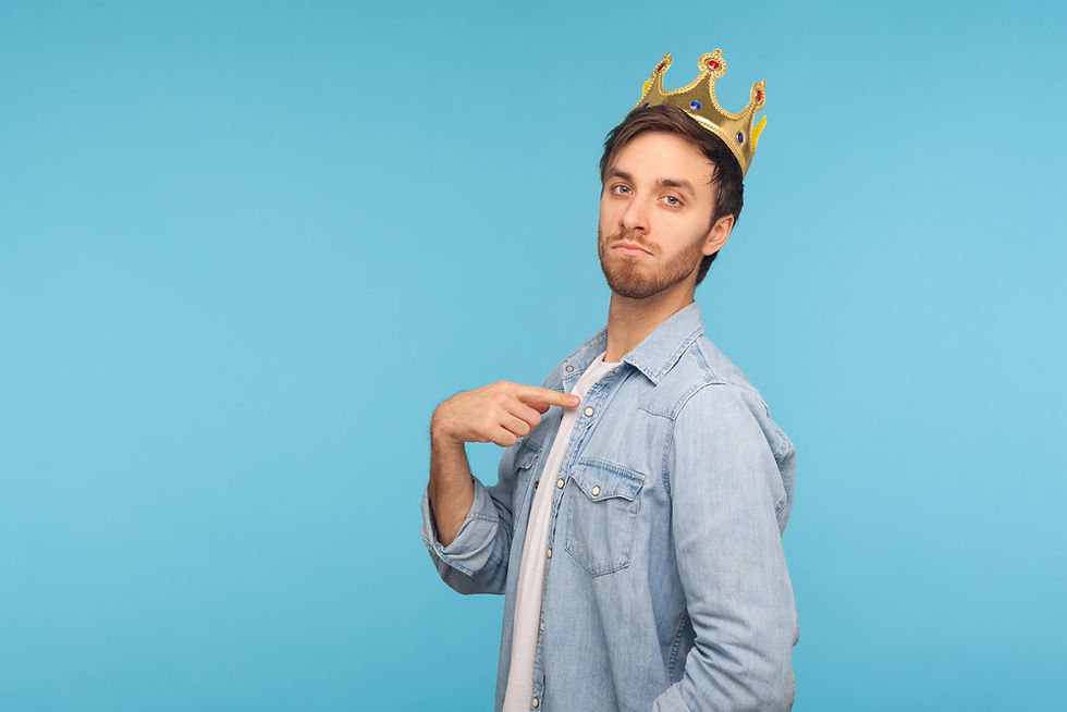 Man in a denim shirt points to himself, wearing a gold crown. Background is solid blue. He has a confident expression.
