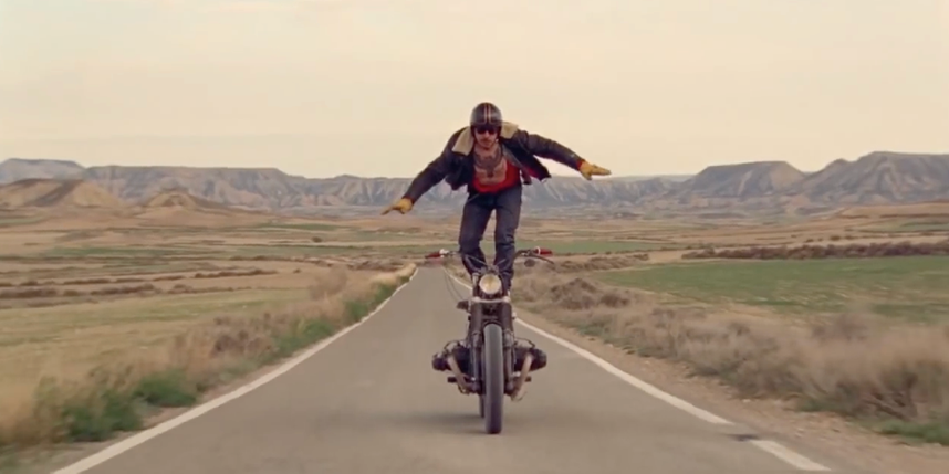 A man standing on top of his motorcycle while it's rolling down a road in the country.