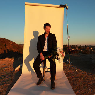 A man sitting on a bar chair, holding a bouquet of flowers, posing outside with a white photography backdrop behind him.