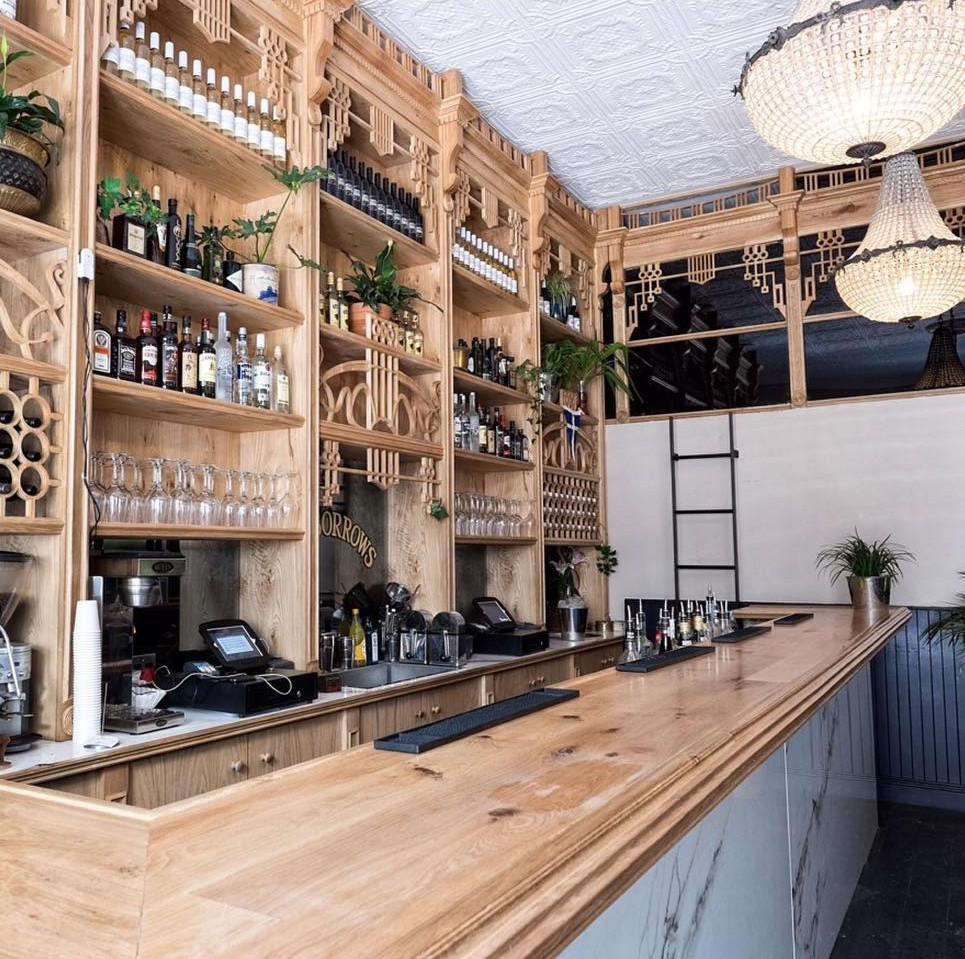 Wide view of The Sorrows bar featuring light oak timber joinery, intricate geometric carvings, built-in shelving stocked with