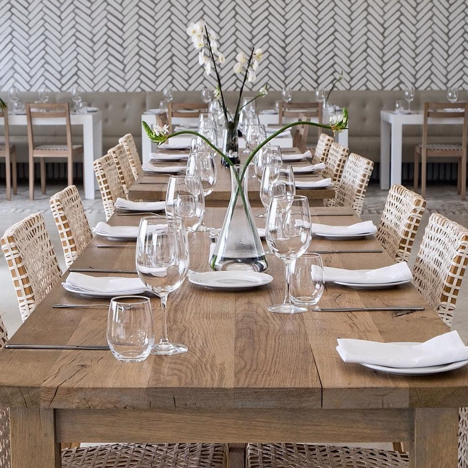 Long wooden dining table set with clear wine glasses, white napkins, and simple silver cutlery, surrounded by woven-back chai