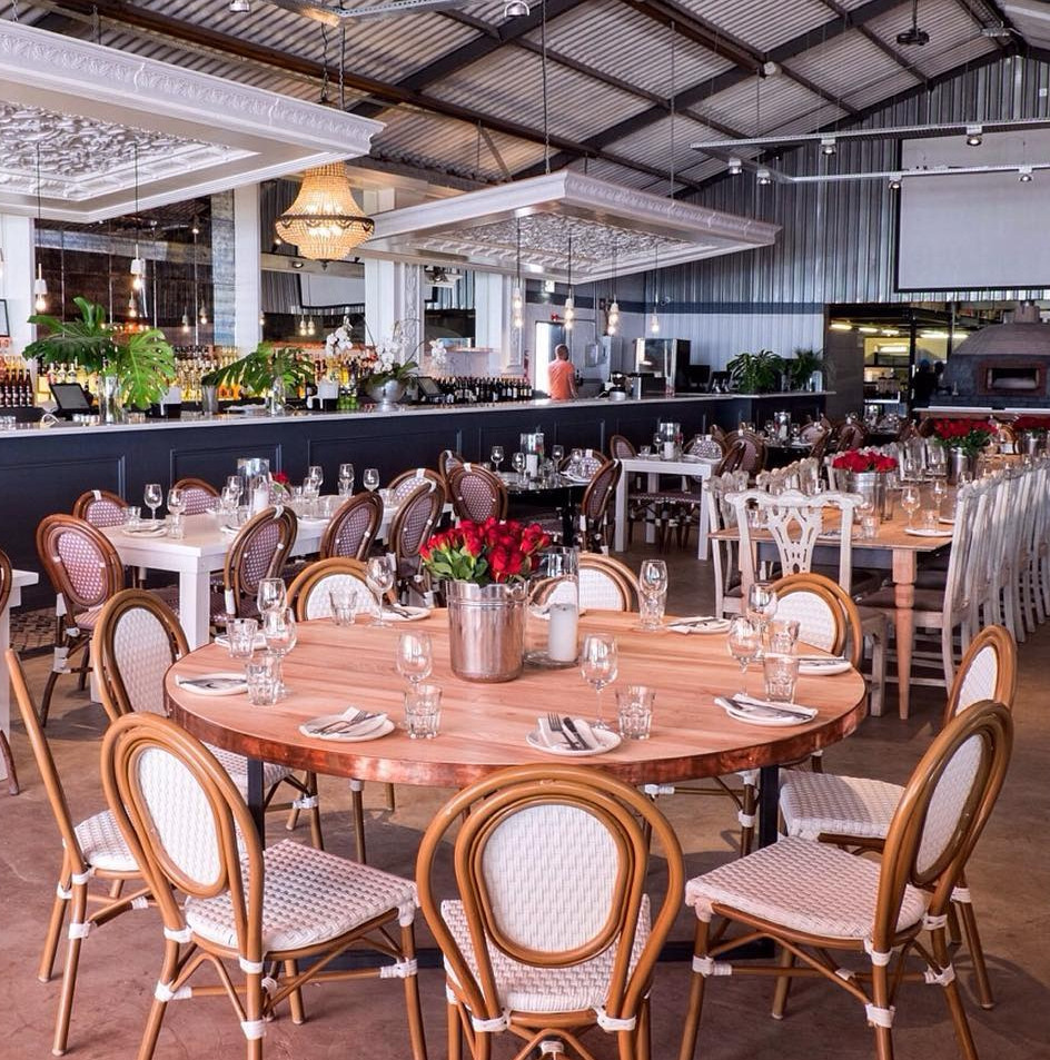 Spacious restaurant interior with round and rectangular wooden tables set for dining, featuring rattan-backed chairs and a vi