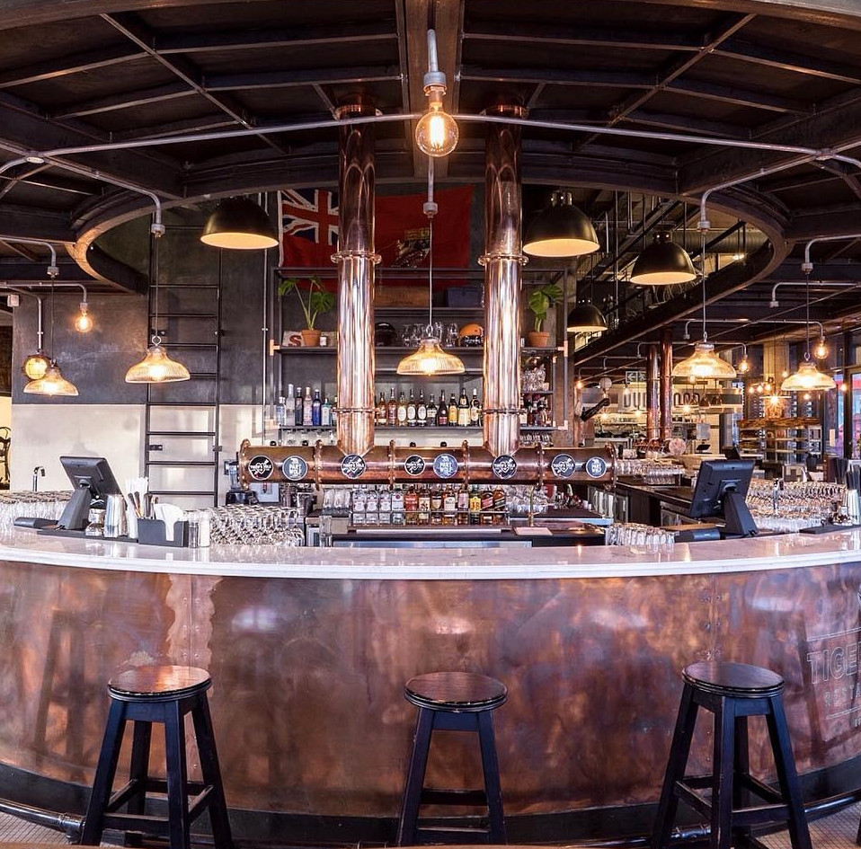 Industrial-style bar interior featuring a curved polished copper counter, black bar stools, overhead pendant lighting, and t