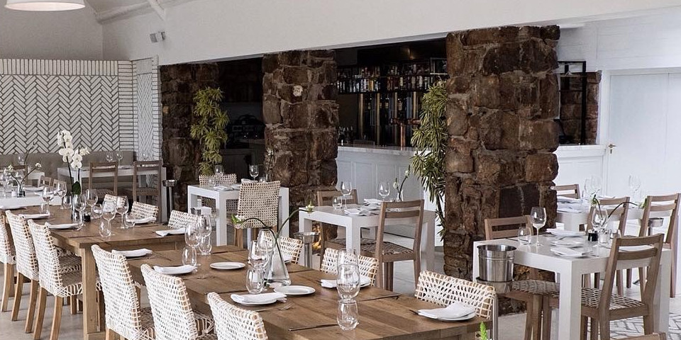 Bright dining space with a mix of long wooden tables and white square tables, stone columns, woven chairs, and a bar area in