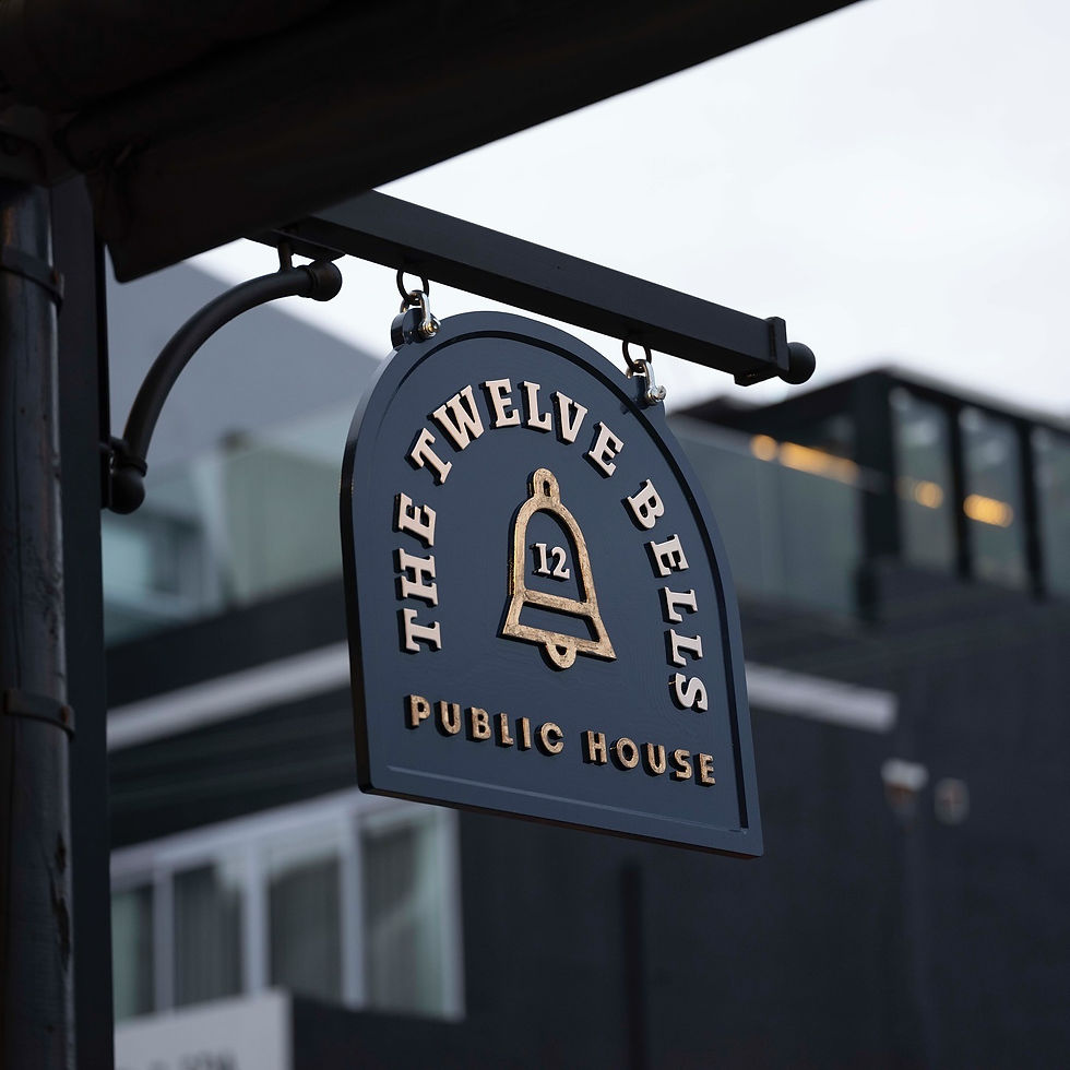 Black and gold hanging sign reading “The Twelve Bells Public House” with a bell illustration, mounted outside the building ag