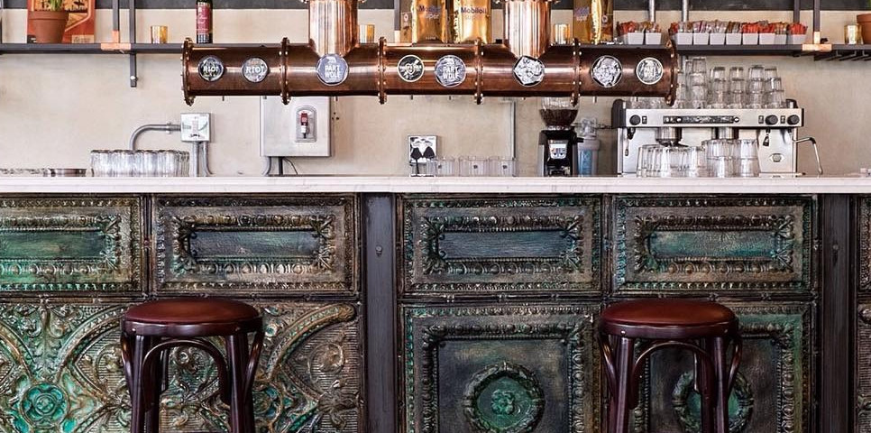 Rustic bar counter with distressed green patina paneling, dark wood bar stools, and a copper beer tap installation above a wh