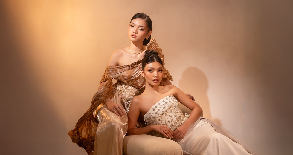 Studio fashion image of two women in formal gowns, one seated and one standing behind, against a warm neutral background.