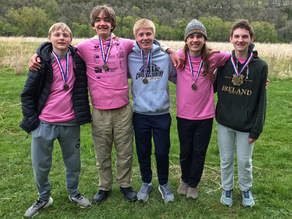 Five smiling teen boys with medals stand arm-in-arm on a grassy field, wearing pink shirts and hoodies, with trees behind.