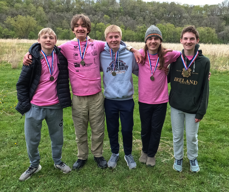Five smiling teen boys with medals stand arm-in-arm on a grassy field, wearing pink shirts and hoodies, with trees behind.