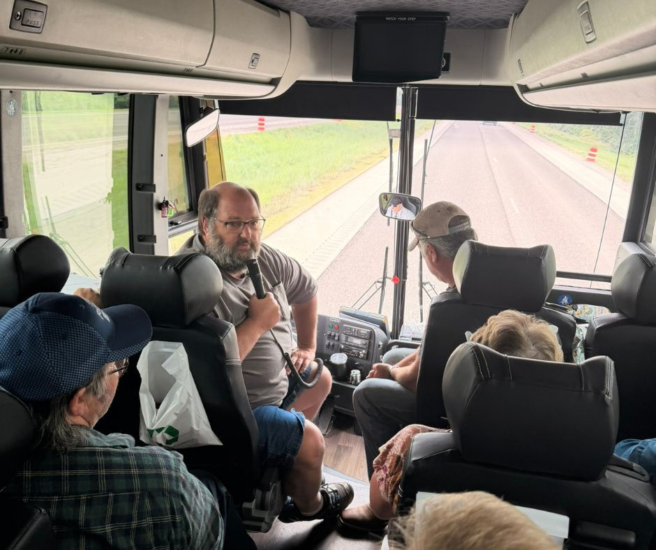 On a bus, a bearded man speaks into a microphone while passengers listen, with a highway stretching ahead.
