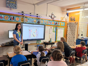 Outreach coordinator Teresa DeMars teaches students in a bright classroom; a screen shows Plant a Pocket of Prairie, with posters, desks, and a calm learning mood.
