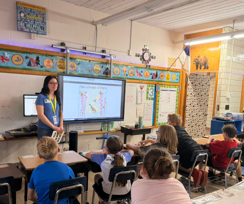 Outreach coordinator Teresa DeMars teaches students in a bright classroom; a screen shows Plant a Pocket of Prairie, with posters, desks, and a calm learning mood.