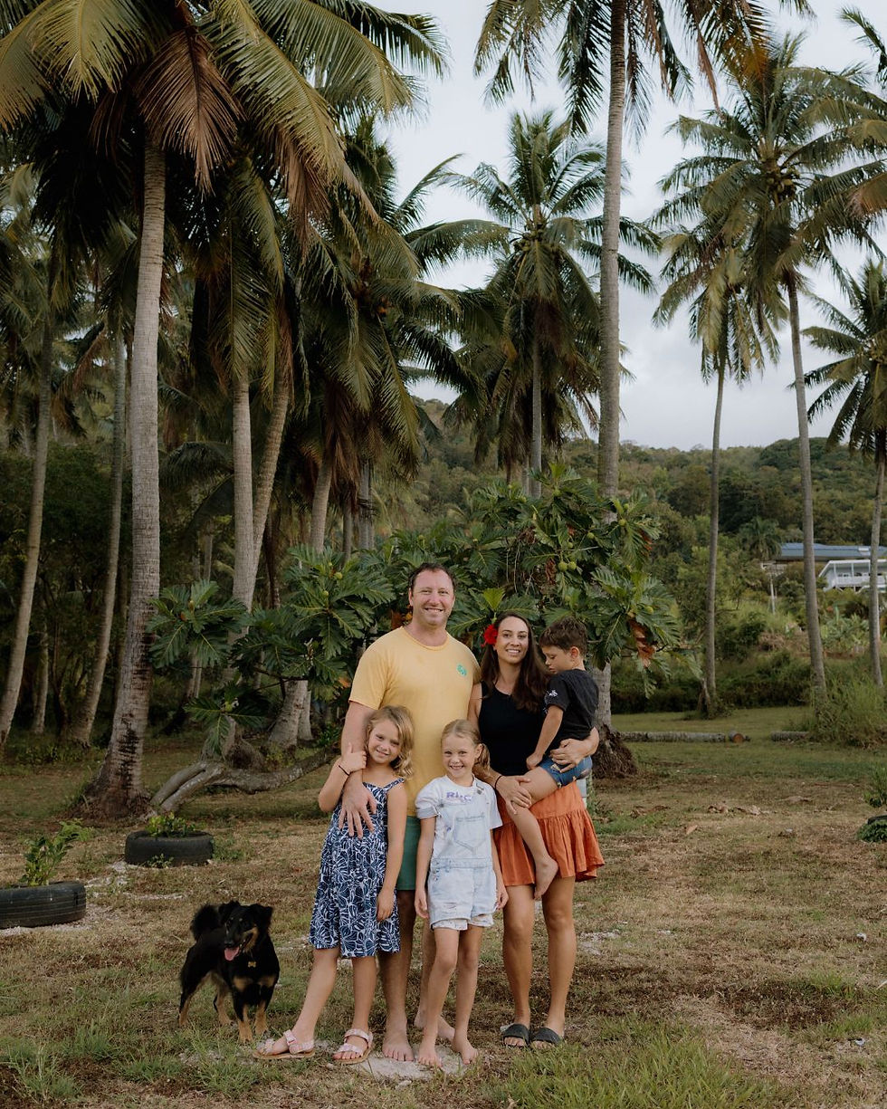 Family of four and a dog in a tropical setting with palm trees. Parents and children smiling, casual attire, relaxed and happy mood.