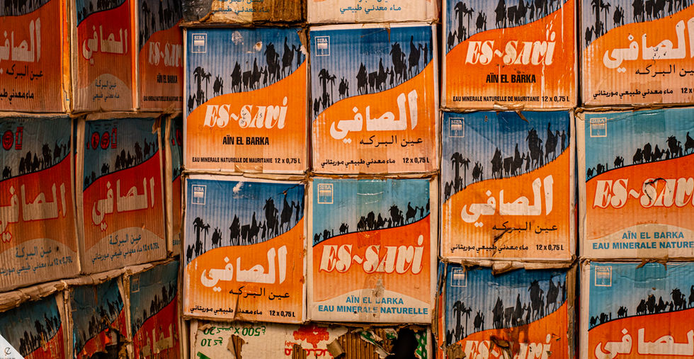 Old boxes of water stacked up on a wall in contrasting colors of blue and orange with a caravan of silhouetted camels walking around the box.