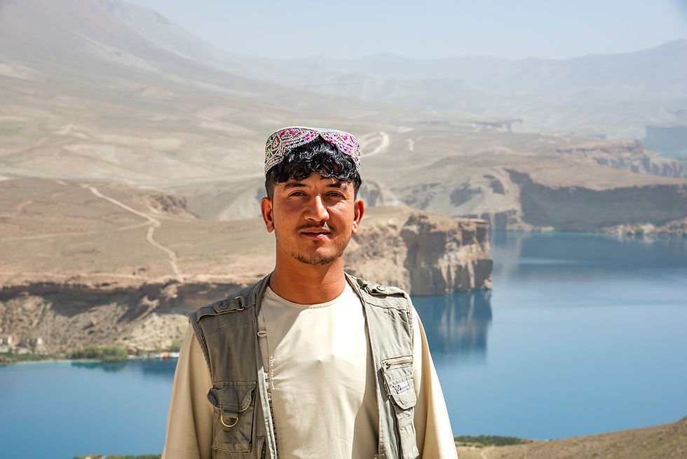 An Afghan man wears a khandahari cap and stands in front of a brilliant blue Band-e Amir Lake in Afghanistan. The surrounding tan-colored hills and cliffs are dull and dry, but the lake looks like a jewel in comparison.