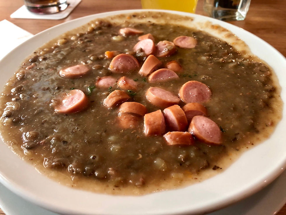 Bowl of lentil soup topped with sausage slices, on a wooden table. A glass of orange juice is visible in the background.