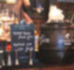 Bar setting with Sipsmith gin barrel, bottles, and a chalkboard listing "Mulled Apple Juice £3.5" and "Mulled Sloe Gin £7.95". Warm ambiance.