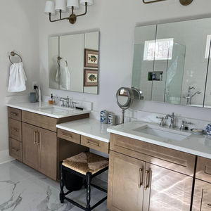 Modern primary bathroom with double vanity, marble countertops, gold hardware, and large mirrors beneath white shaded sconces.