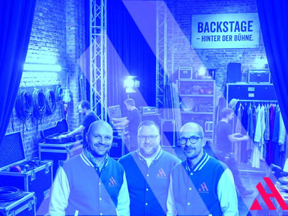 Drei lächelnde Männer der APARTMENTHELDEN in blau-weißen College-Jacken stehen im Vordergrund eines Backstage-Bereichs mit technischem Equipment und Ziegelwand. Im Hintergrund hängt ein Schild mit der Aufschrift „BACKSTAGE – HINTER DER BÜHNE“. Das Bild hat einen bläulichen Filter und zeigt das Firmenlogo in der unteren rechten Ecke.