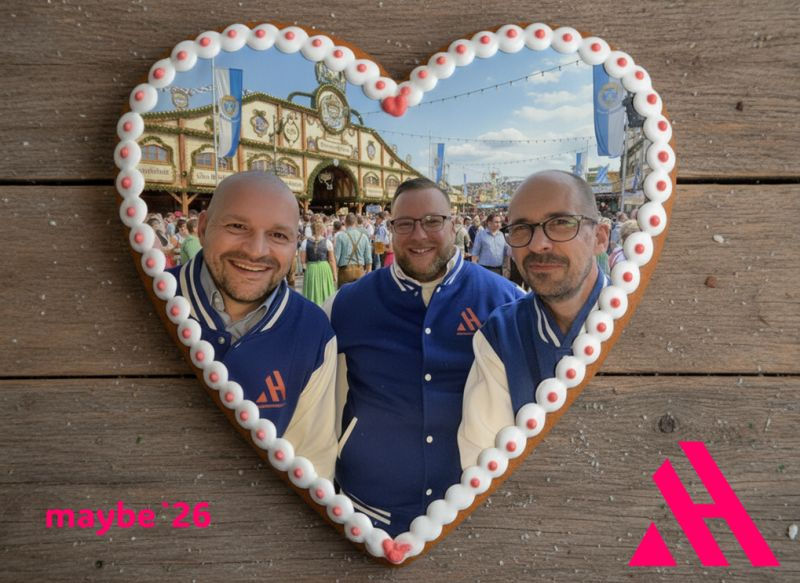 Ein Lebkuchenherz im Oktoberfest-Stil liegt auf einer Holzoberfläche. Im Inneren des Herzens ist ein Foto von den drei APARTMENTHELDEN zu sehen, die auf der Wiesn vor einem Festzelt stehen. Sie tragen blaue College-Jacken mit ihrem Logo und lächeln. Unten rechts im Bild ist das pinke "AH"-Logo der APARTMENTHELDEN platziert.