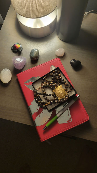 Red leather journal with mala beads, crystals, and pen arranged on a wooden nightstand as part of a spiritual travel routine setup, including amethyst, rose quartz, and other healing stones.