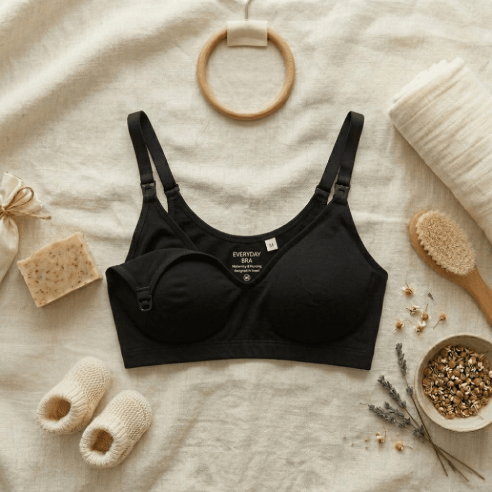 The Yael seamless nursing bra in black by Yael and Michal, flat lay on cream linen with baby booties and natural accessories
