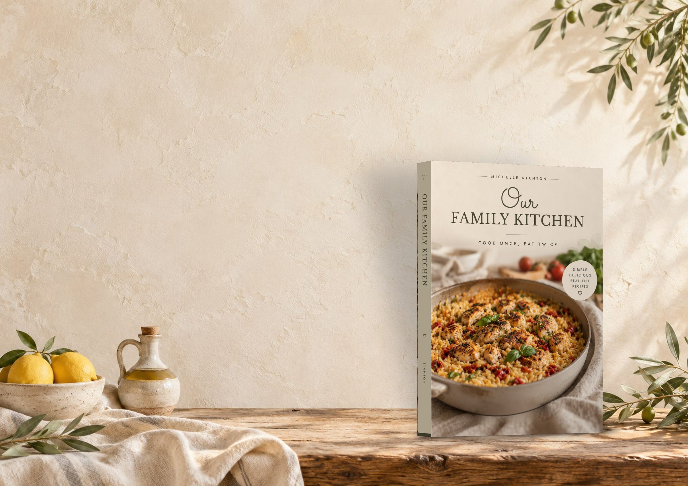 The Our Family Kitchen cookbook standing on a rustic wooden kitchen counter beside lemons and olive branches