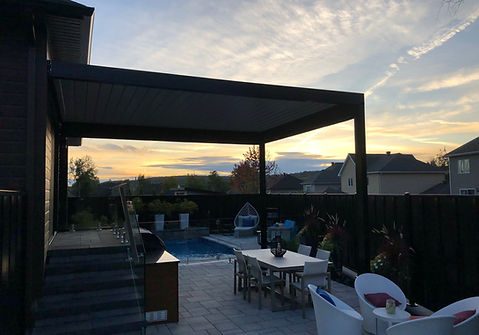 Pergola bioclimatique noire avec lames orientables aménagée au-dessus d’une terrasse extérieure au coucher du soleil