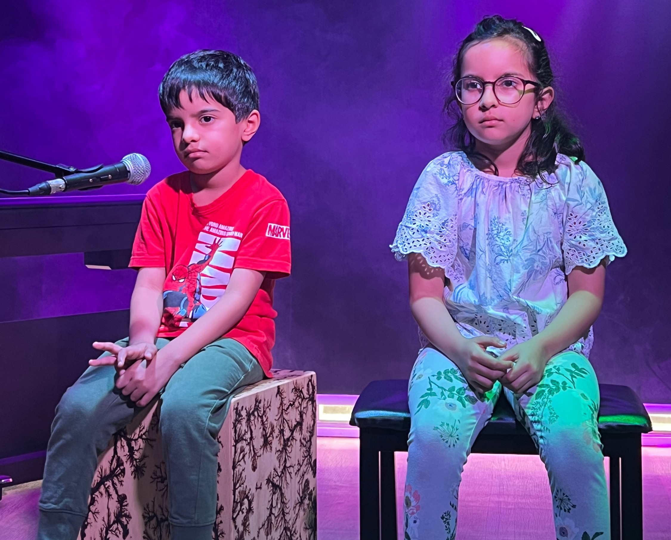 Two solemn children sit on stools on a purple-lit stage beside a microphone; boy’s red MARVEL shirt stands out.
