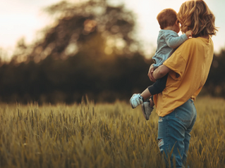 a mother in a yellow jumper holding a toddler whilst standing in a field at golden hour