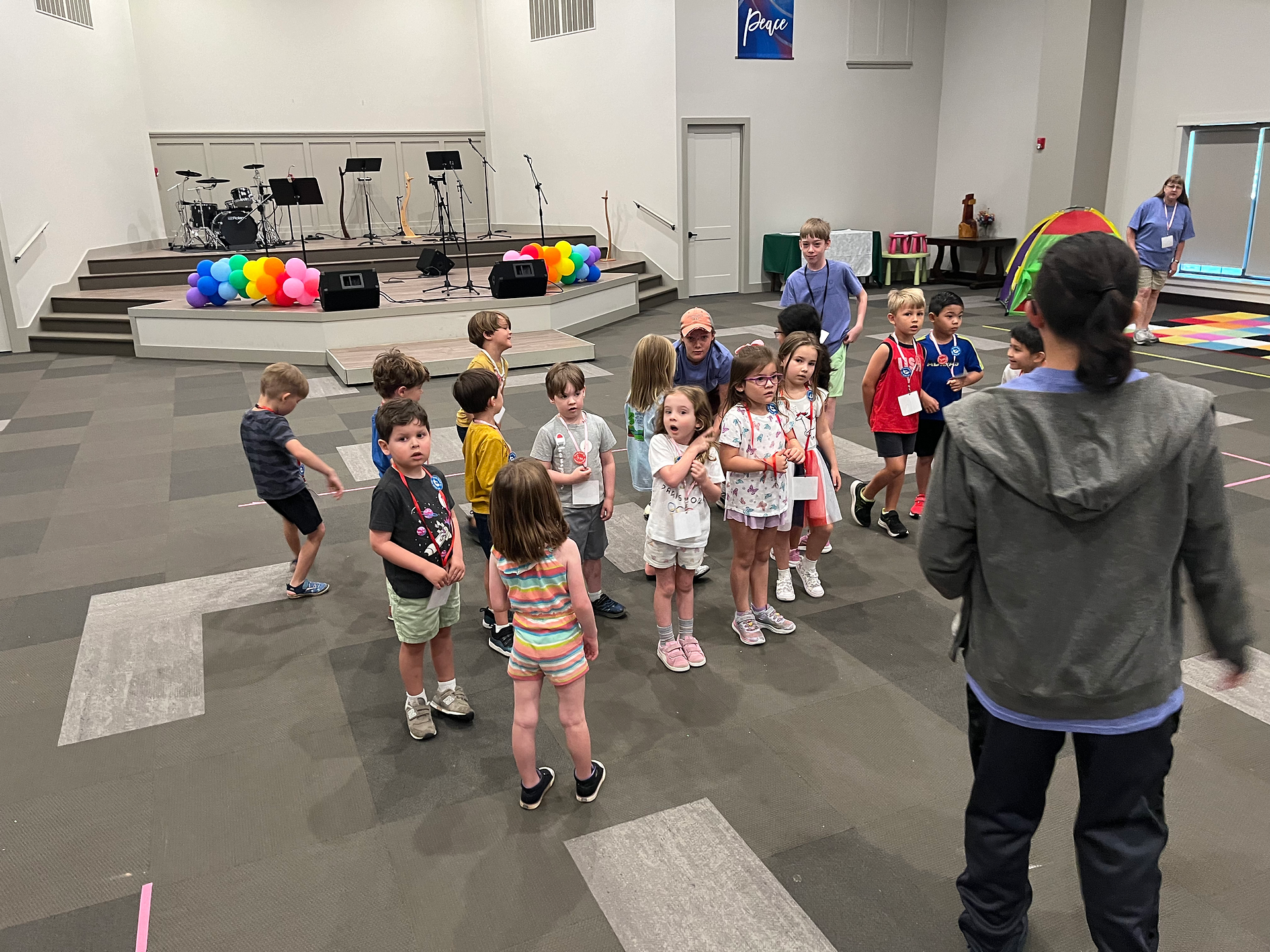 A group of young children stand listening to and looking at a teen boy in a  hoodie in front of them.