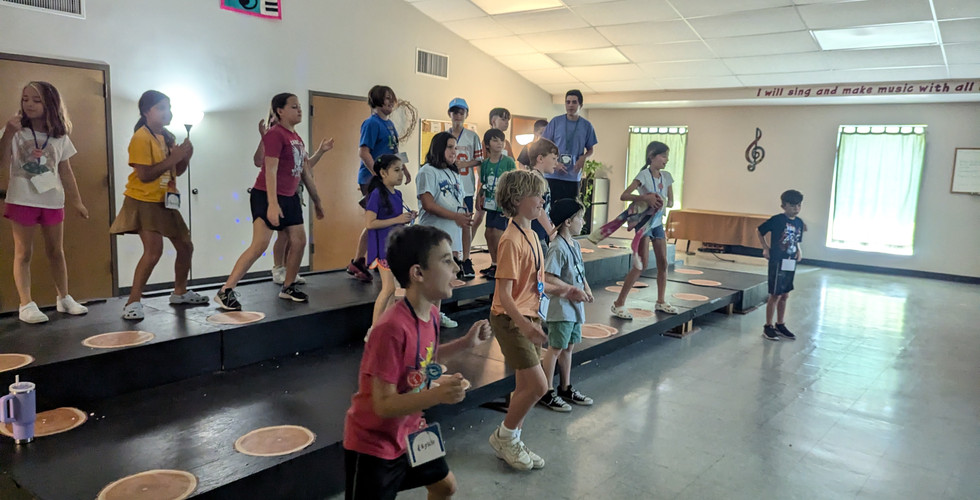 A group of children on black risers are in the middle of a dance with their arms up.