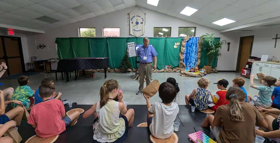 A group of children are sitting on black risers looking ahead at an older man standing in front of them holding a piece of paper with the words "Wesley Grace" on it.
